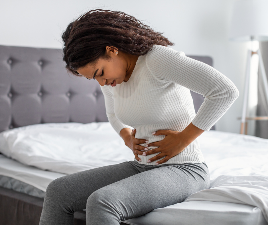 Woman who appears to be hunched forward in pain, is sitting on a bed and holding her hands to her stomach.