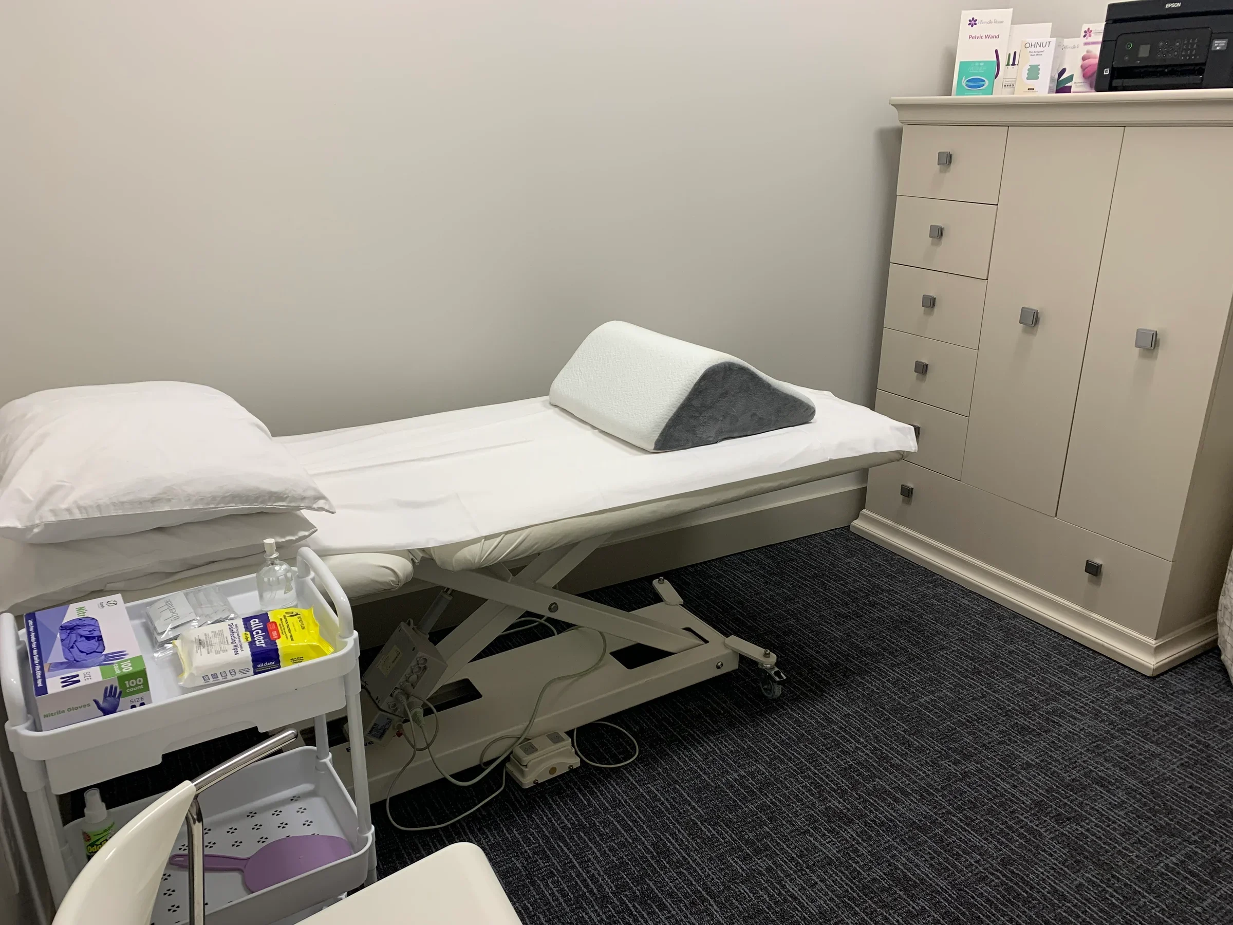 Medical examination room with an adjustable bed, pillow, blanket, medical supplies on a cart, and a cabinet with informational brochures and equipment.