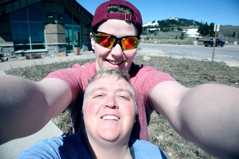 Two smiling people taking a selfie outdoors on a sunny day with a building in the background.