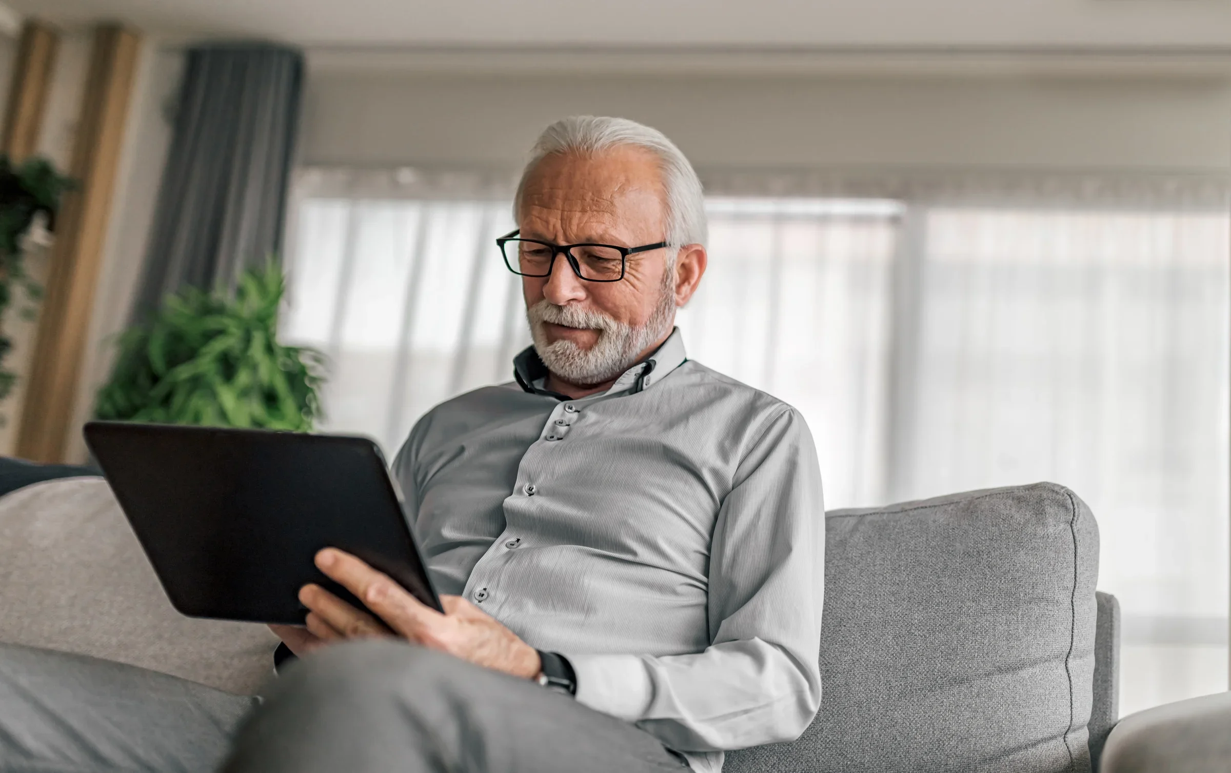 An elderly man with white hair, glasses, and a beard, sitting on a gray sofa and looking at a black tablet in his hands in a bright living room.