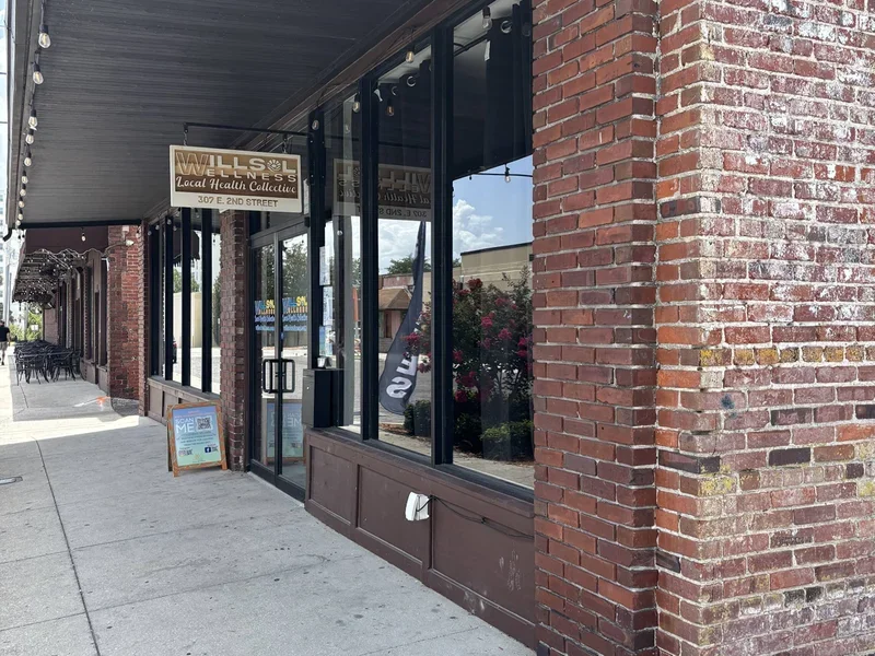 Exterior of a brick storefront with glass windows and a sign that reads "Wells & Co. Local Health Collective." Sidewalk in front and a menu board outside.
