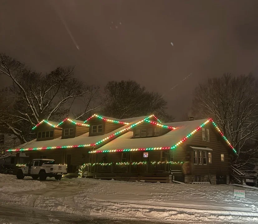 Log Cabin covered in snow with Christmas lights on top roofline and railing