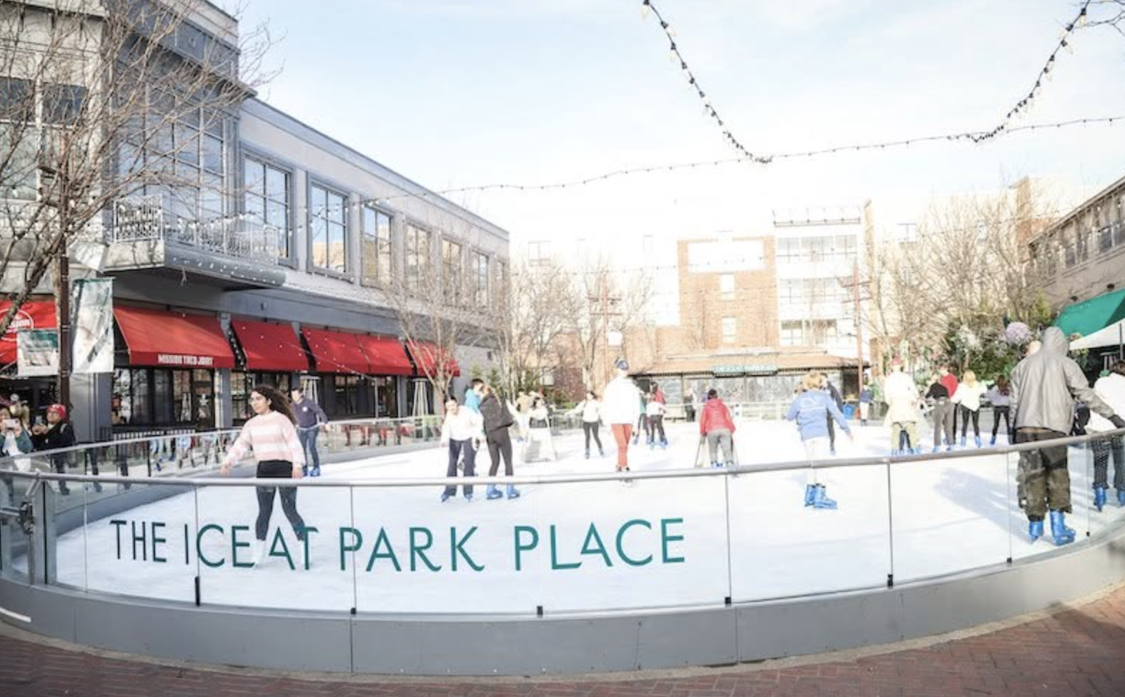 People ice skating outdoors at The Ice at Park Place skating rink, surrounded by buildings and trees.