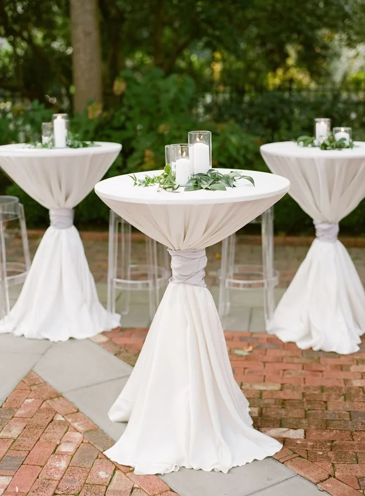 Outdoor celebration tables with white tablecloths, candles, and greenery on a brick and concrete patio with trees in the background.