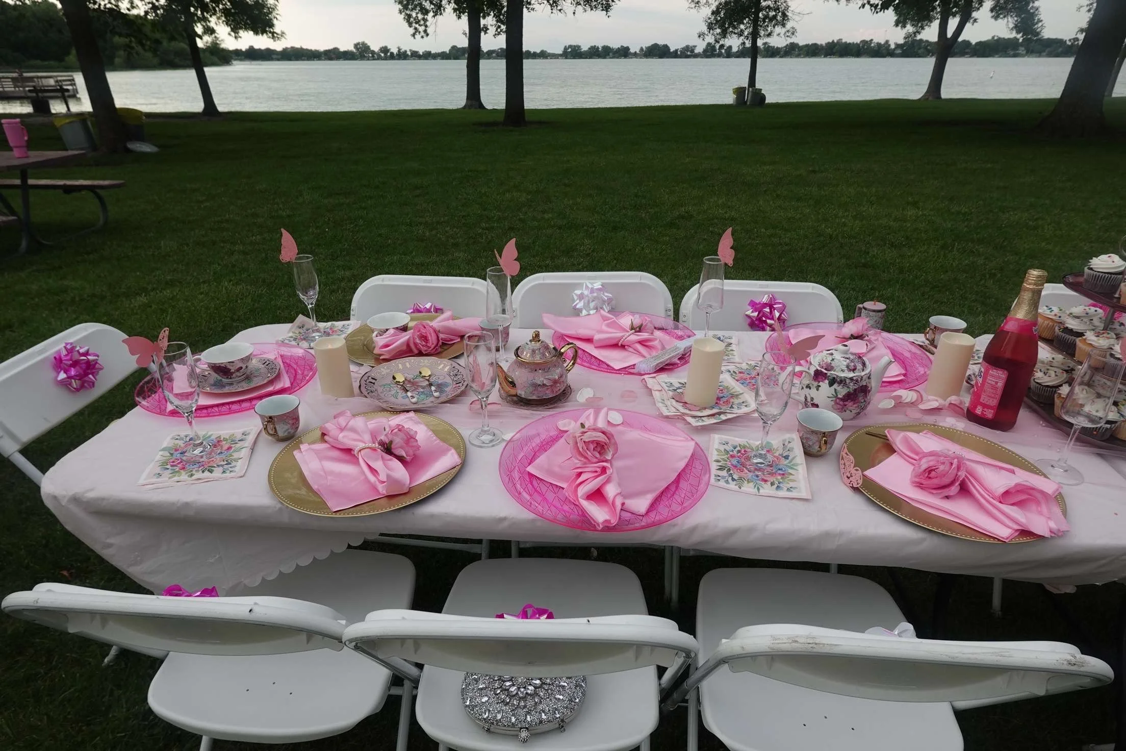 A decorated outdoor table set for a celebration near a lake, with pink napkins, floral tableware, candles, and pink butterfly decorations.