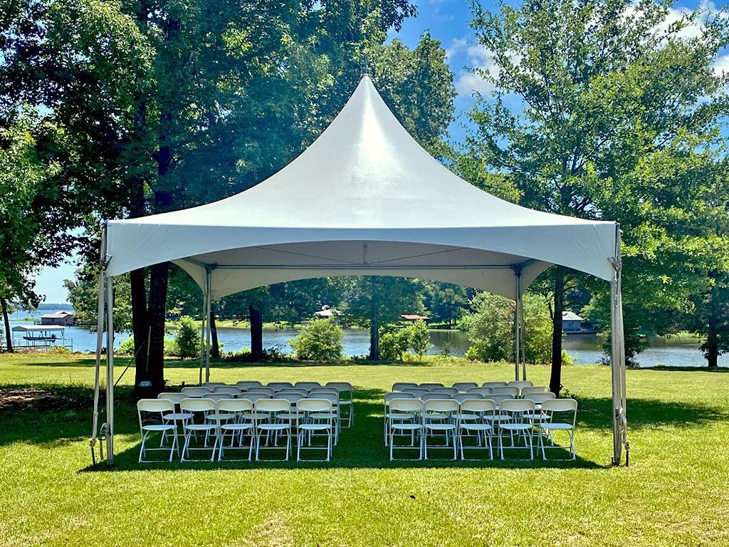 A white outdoor event tent set up on grassy ground with folding chairs arranged inside, overlooking a lake with trees and a dock in the background.