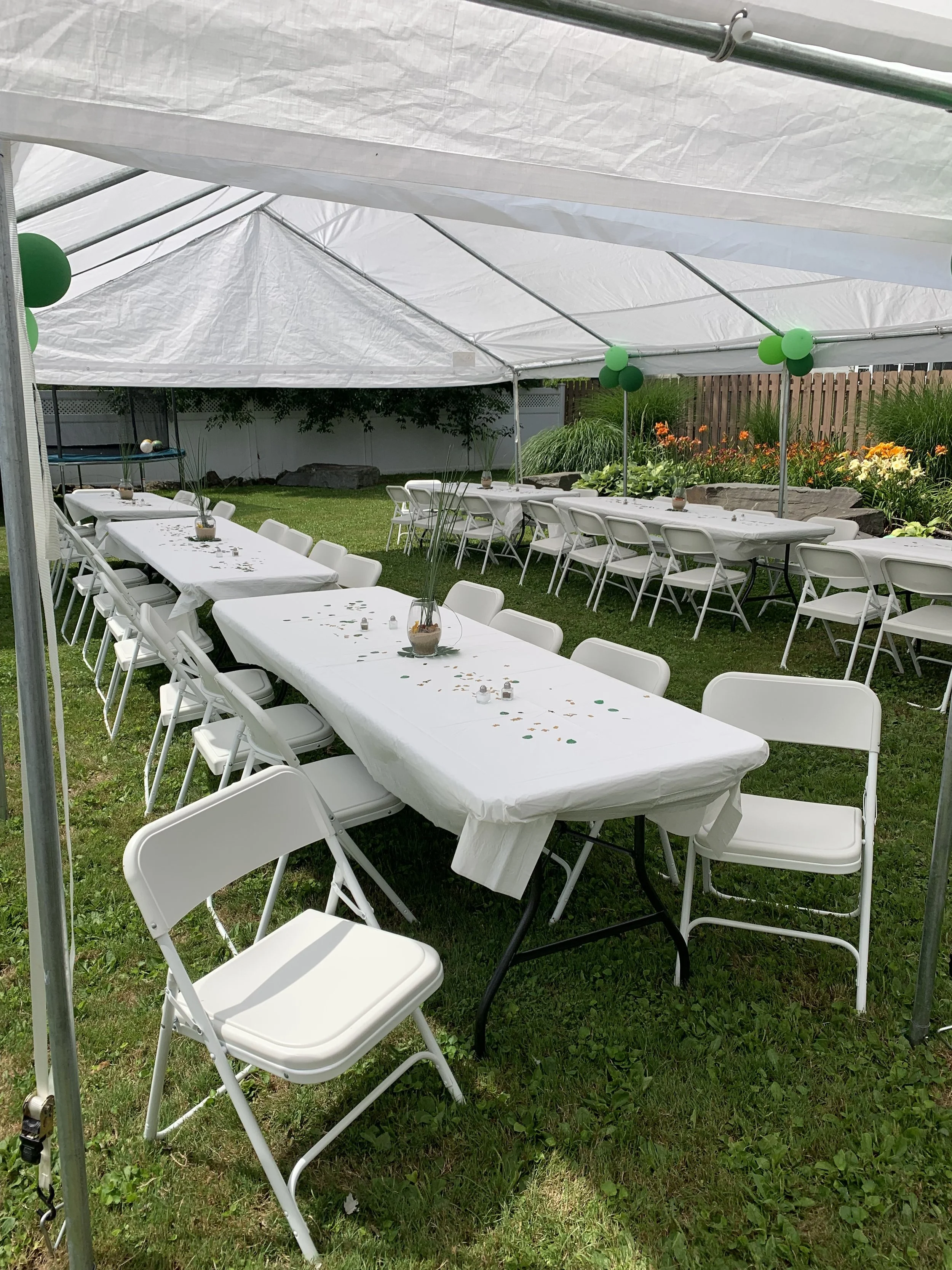 Party setup in a backyard with long tables covered in white tablecloths, surrounded by white folding chairs, under a white canopy with green balloons for decoration, and a flower garden in the background.