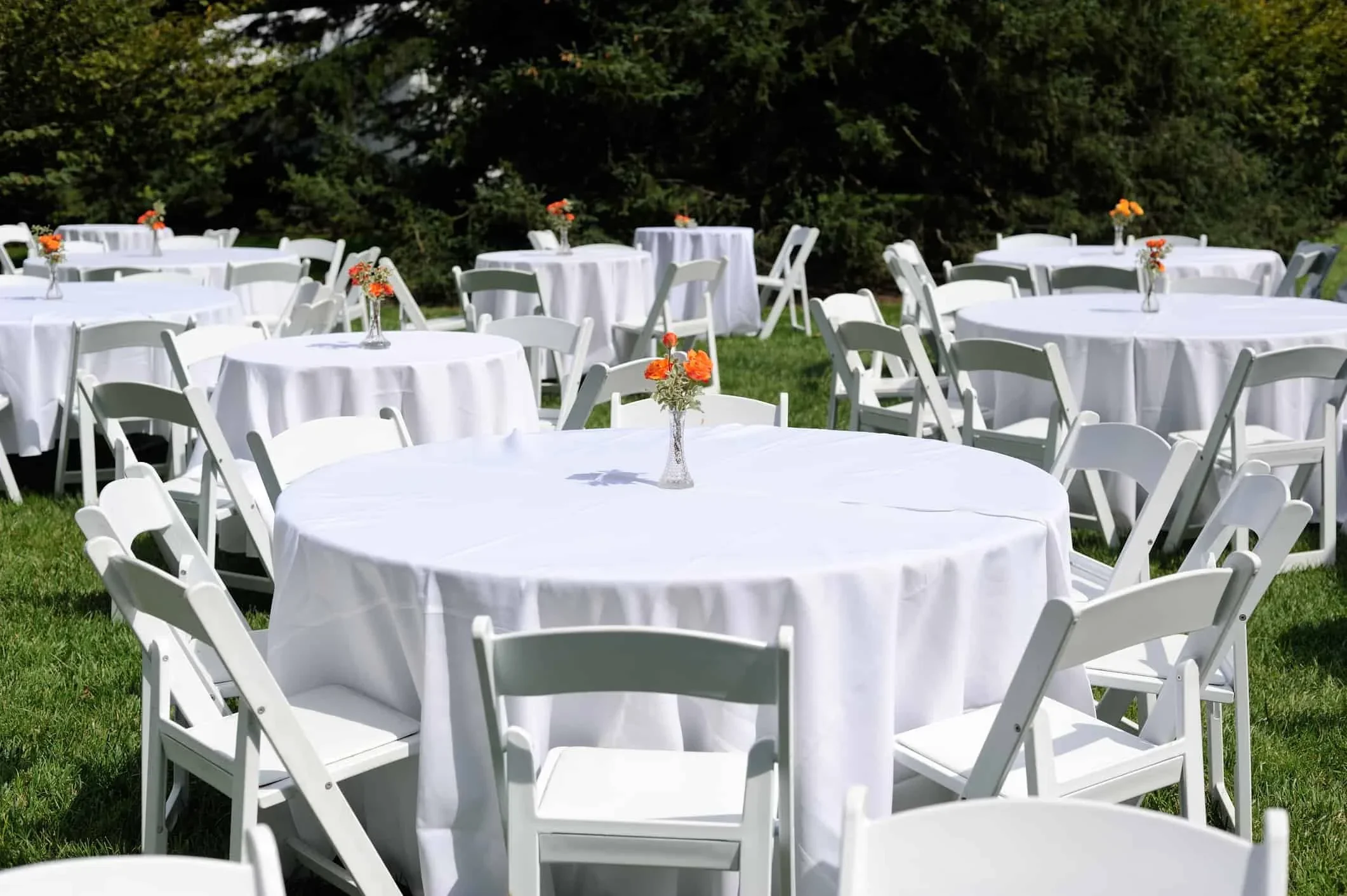 Outdoor event setup with round tables covered in white tablecloths and small flower arrangements in glass vases on a grassy area with trees in the background.