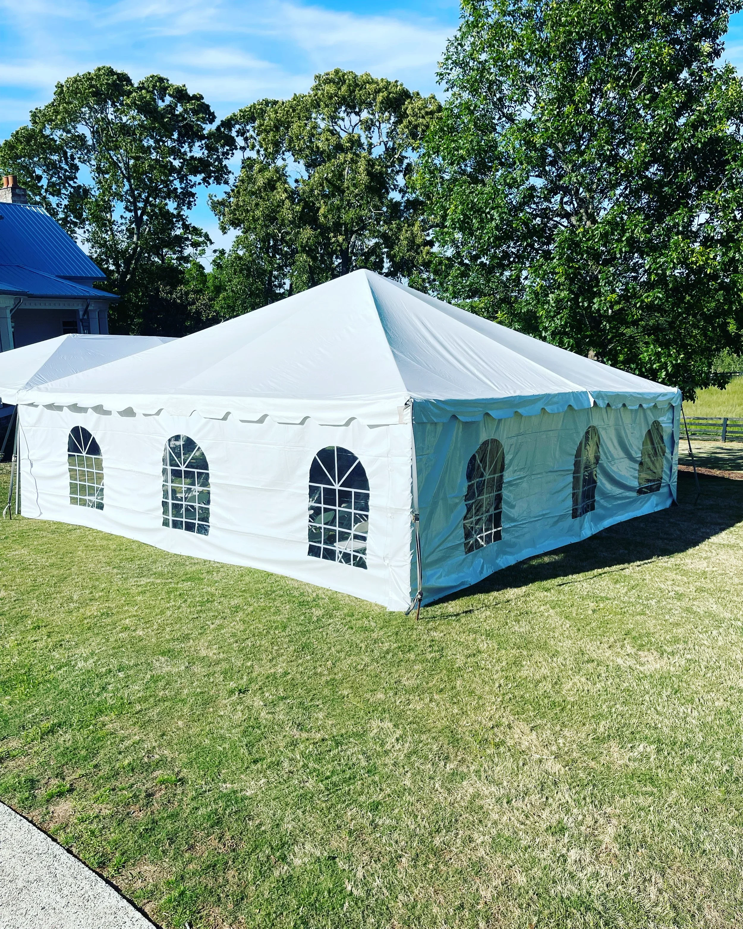 White outdoor event tent with arched window panels on a grassy area, surrounded by trees and a blue sky.