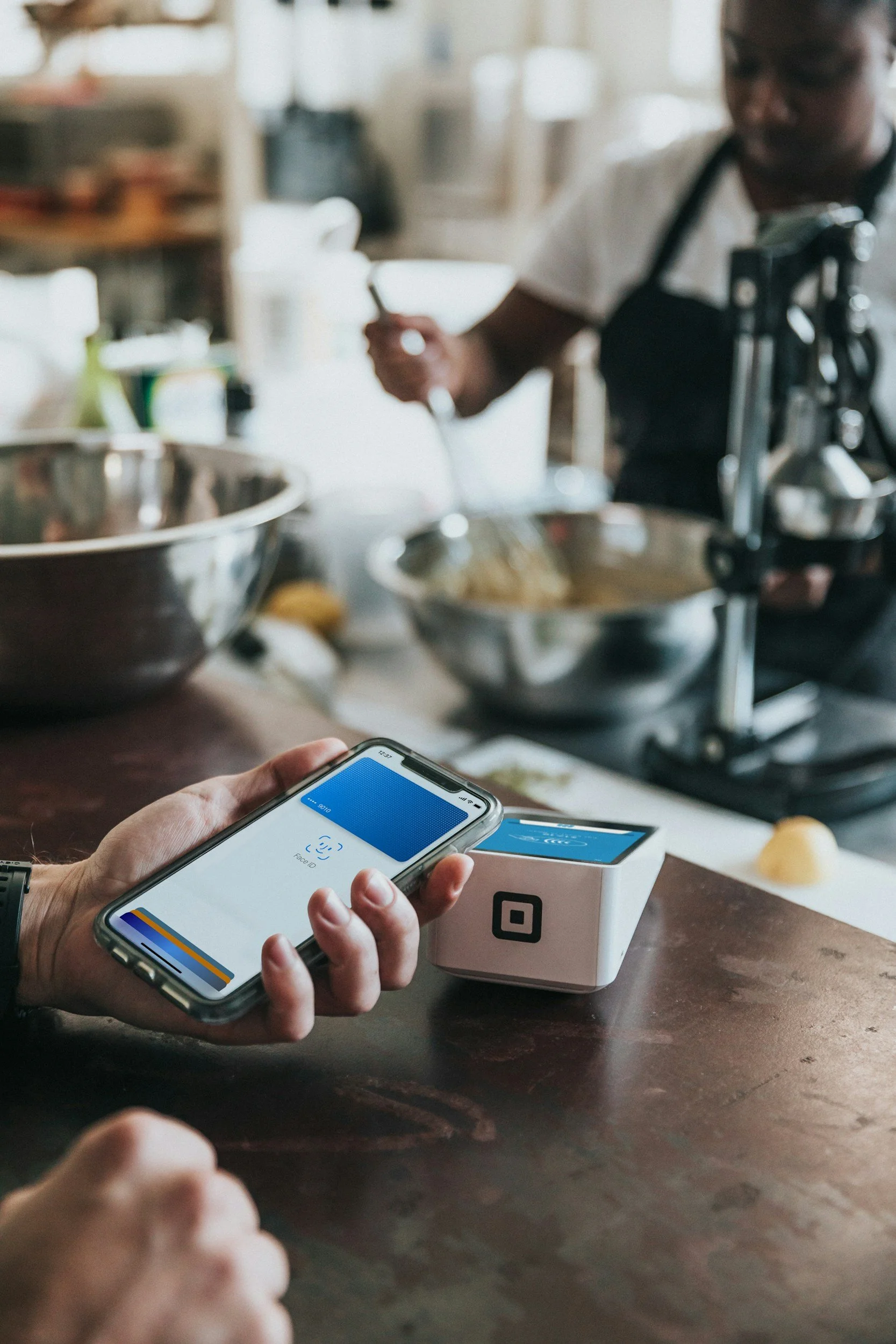 Person using a smartphone with a contactless payment app in a kitchen or bakery, with a baker mixing ingredients in the background.