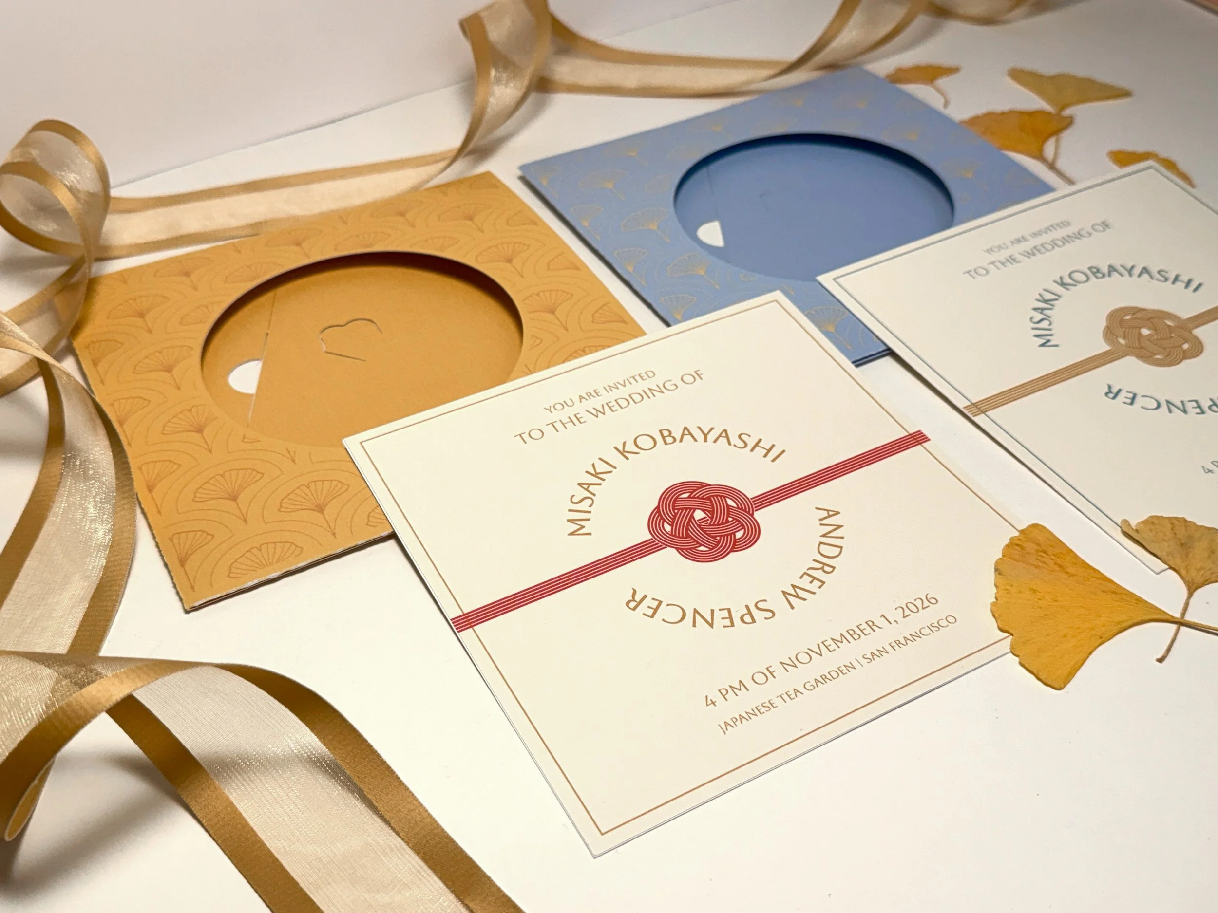 Wedding invitation card with an elegant white background, gold and blue decorative overlay, and dried ginkgo leaves and ribbons around it.