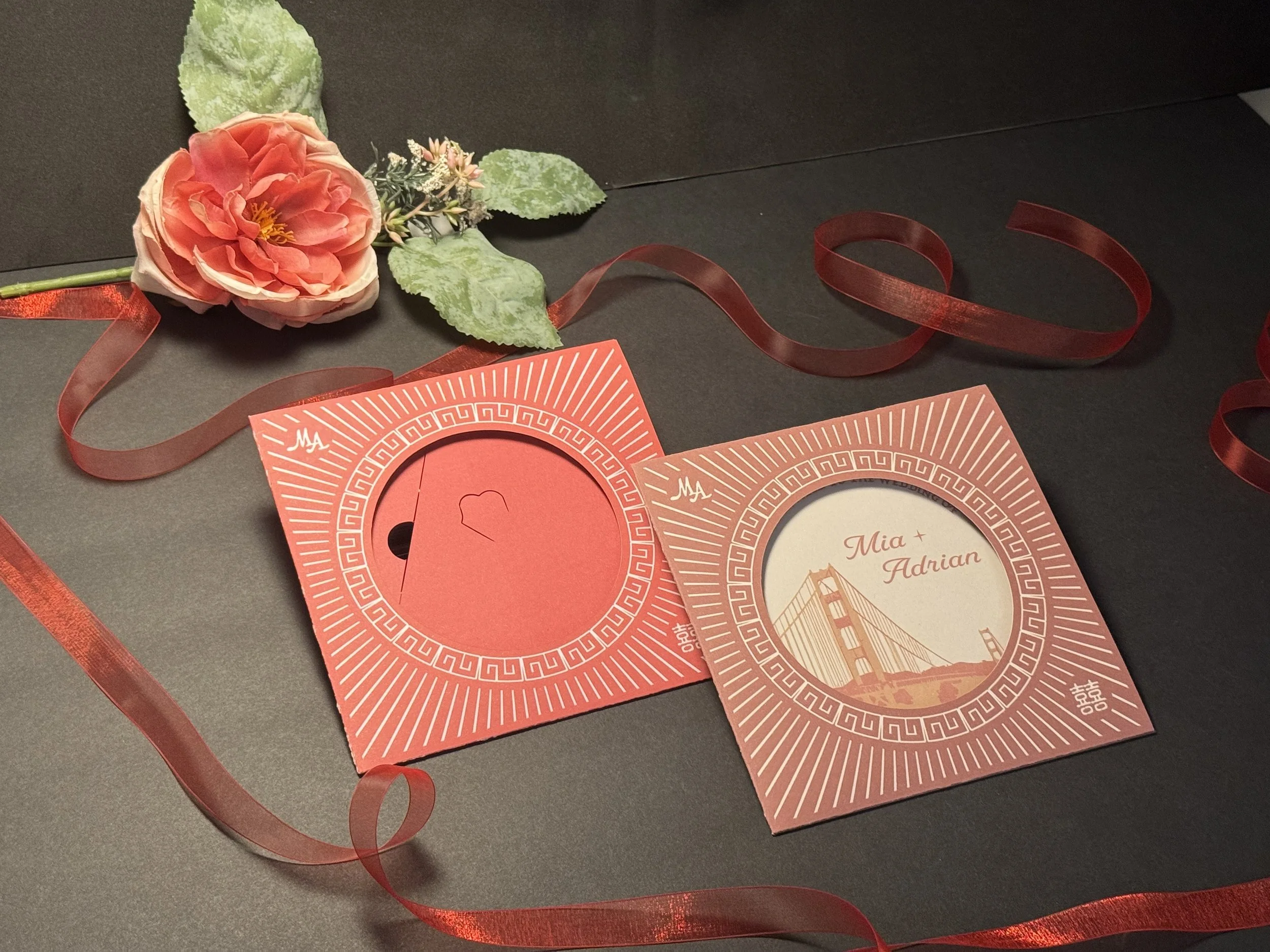 A black surface with a Nostalgic wedding invitation set or engagement invitation cards paired with a pink flower with green leaves, surrounded by red ribbon.