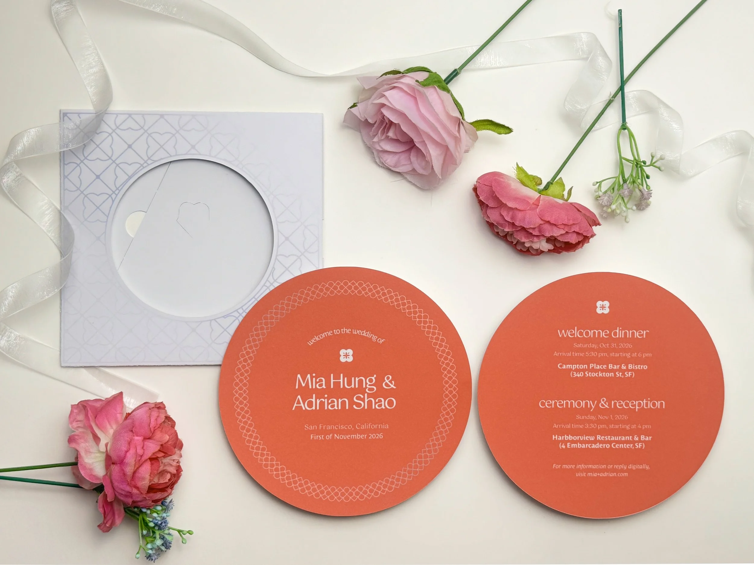 Wedding invitation cards, pink artificial flowers, and white decorative ribbon on a white surface.