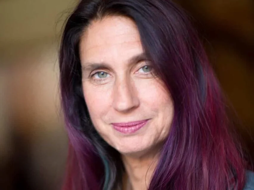 Close-up portrait of a woman with long, straight dark hair featuring vibrant purple highlights, light green eyes, and a soft, neutral expression.