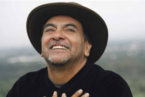 Smiling hispanic man who is don Miguel Ruiz, in hat, with hands on chest