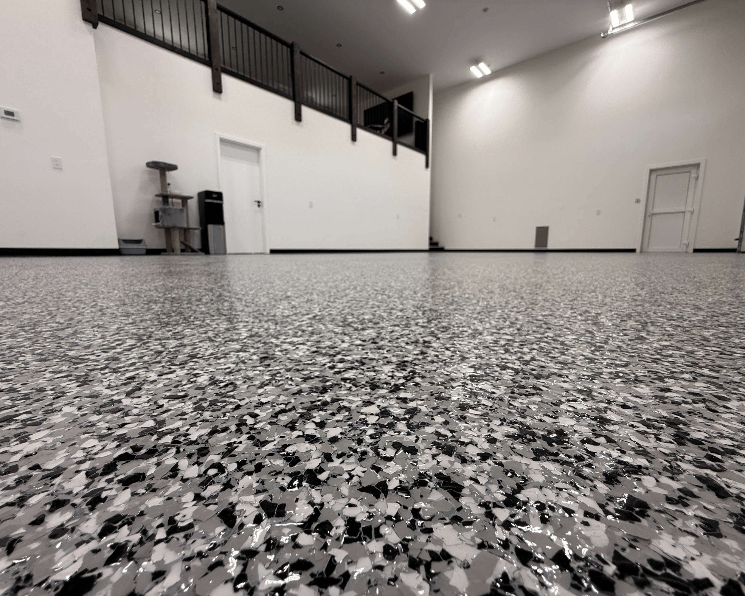 Empty room with speckled black, white, and gray terrazzo flooring, white walls, and a white door. There is black railing on an upper level and minimal furniture including a small table and a water dispenser.