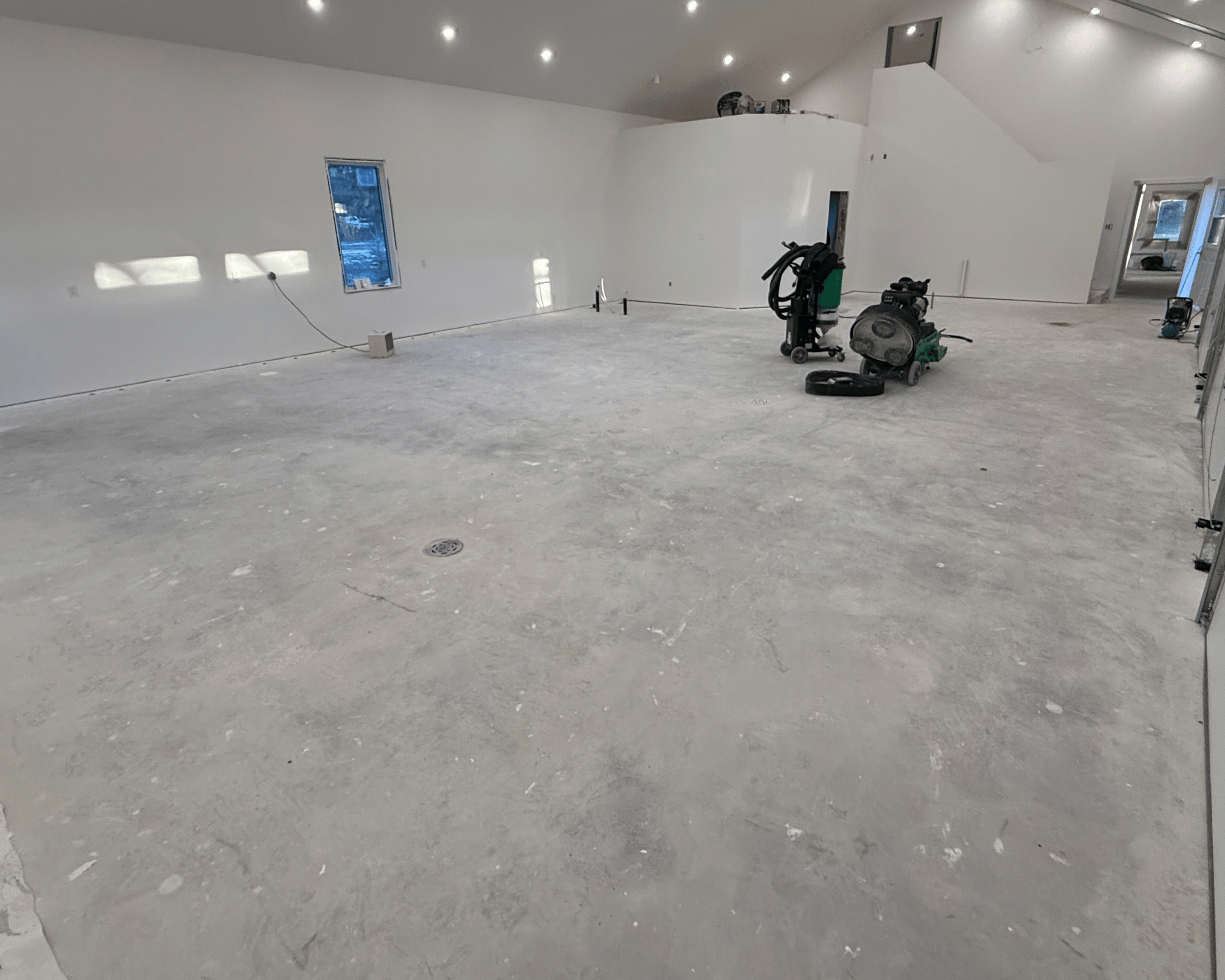 Construction site of an interior room with unfinished concrete floor, white walls, small window, and construction equipment.