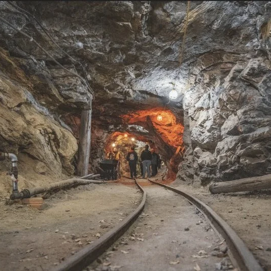People walking inside a tunnel with train tracks, illuminated by lights with a rocky ceiling and walls.