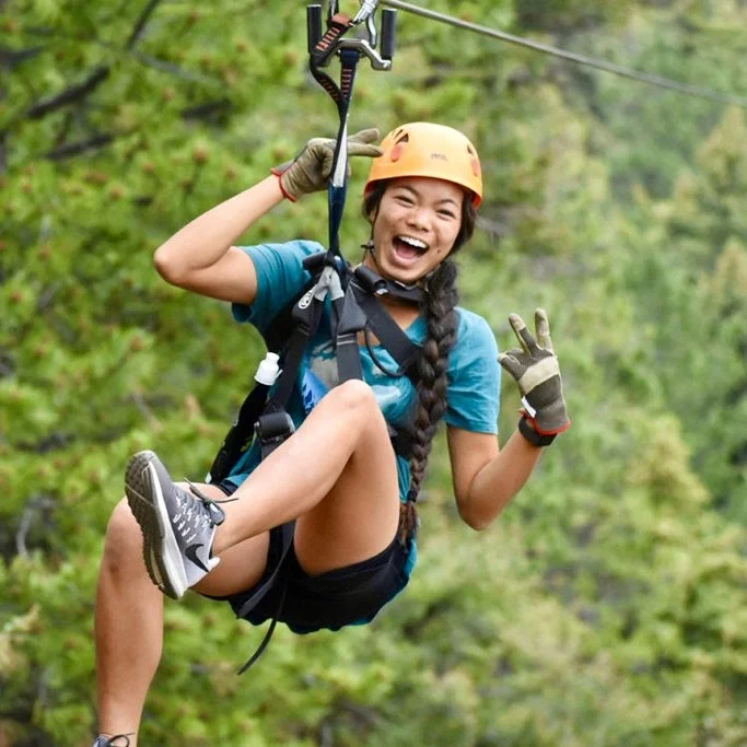 Young woman with long braided hair ziplining, smiling, wearing a helmet, gloves, athletic clothes, and sneakers, in a forested area.