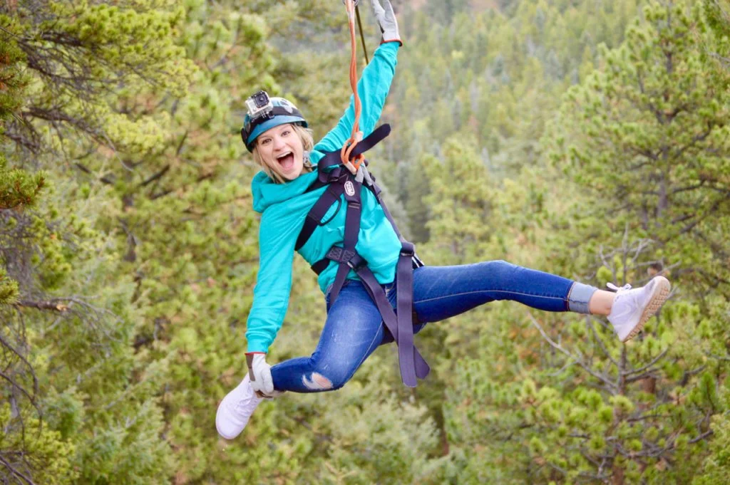 Girl zip-lining through a forested area, smiling excitedly, wearing a helmet and harness.