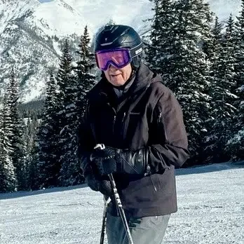 A person in winter gear and a helmet holding ski poles in a snowy mountain landscape with evergreen trees.