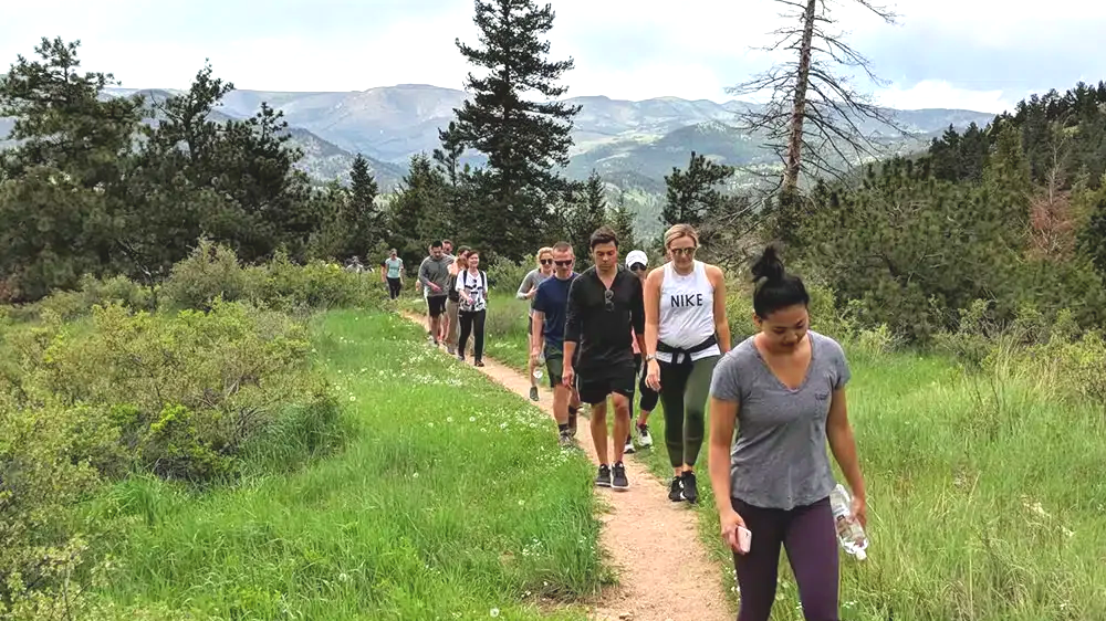 A group of people walking on a dirt trail through a green, hilly landscape with trees and mountains in the background.