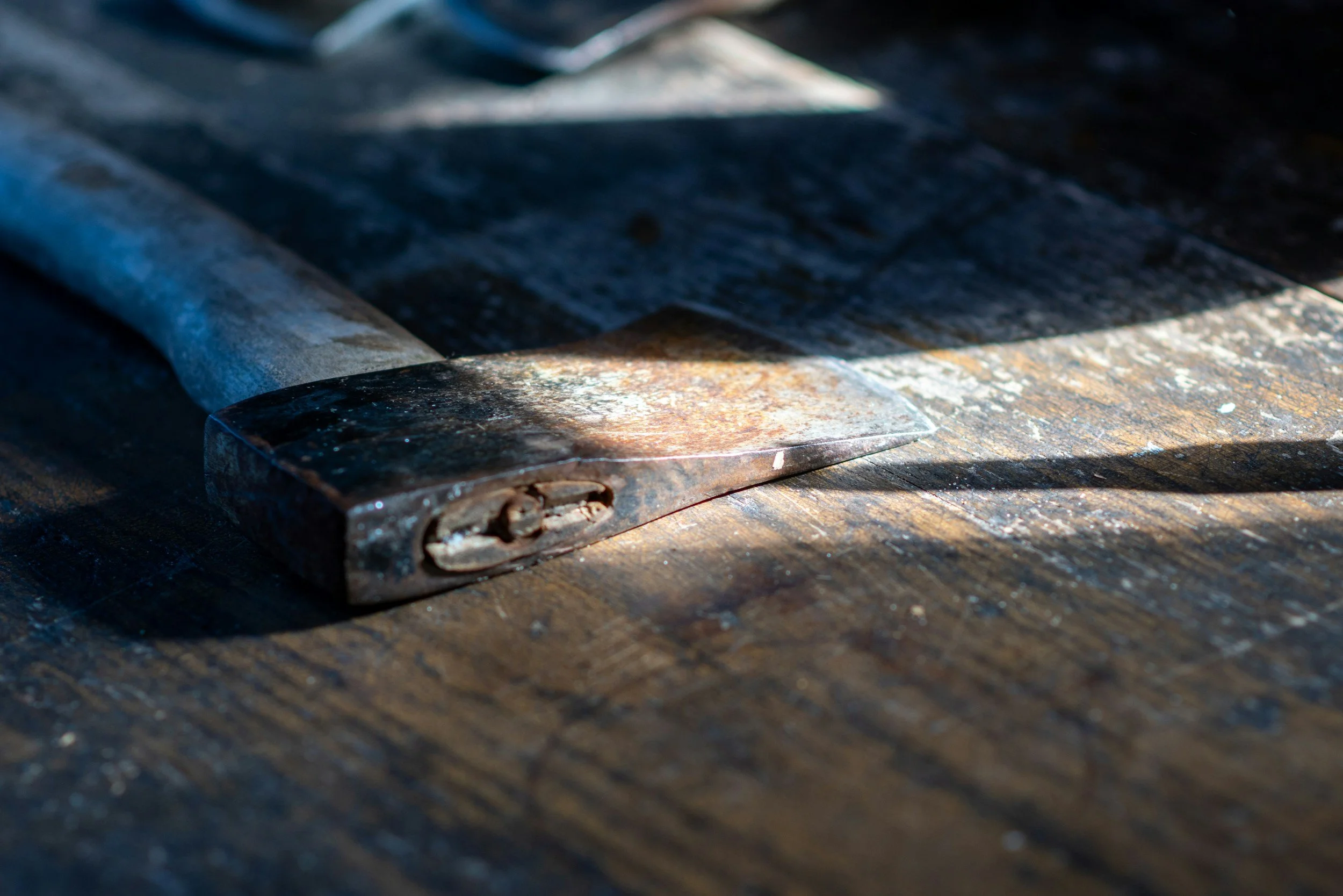 Close-up of a rusty, old, curved axe head resting on a worn wooden surface with shadows.