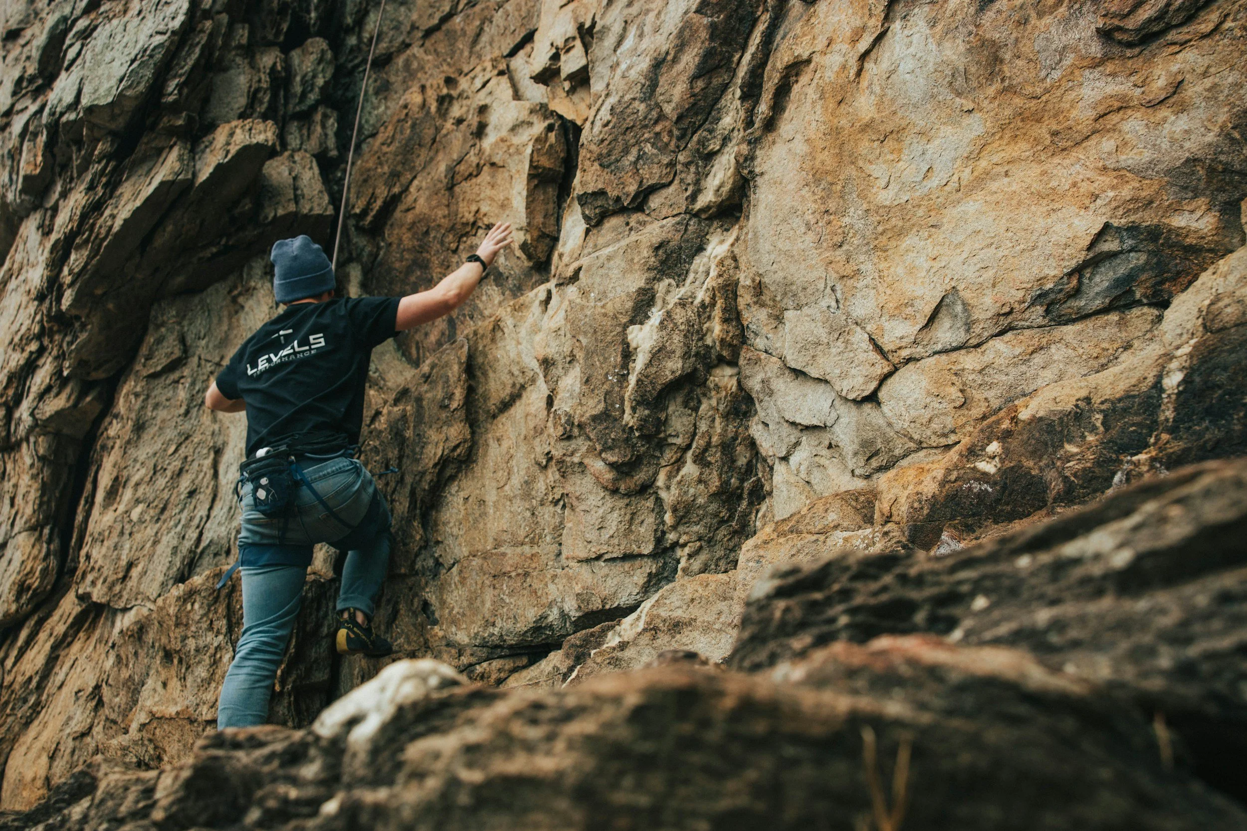 A person wearing a blue hat and climbing gear is rock climbing on a rugged cliff face.