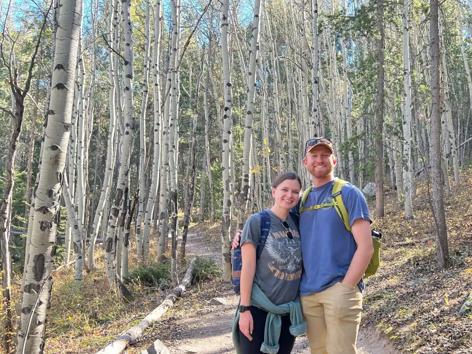 A smiling couple stands on a dirt trail in a forest of tall aspen trees, dressed in casual outdoor clothing and carrying backpacks.