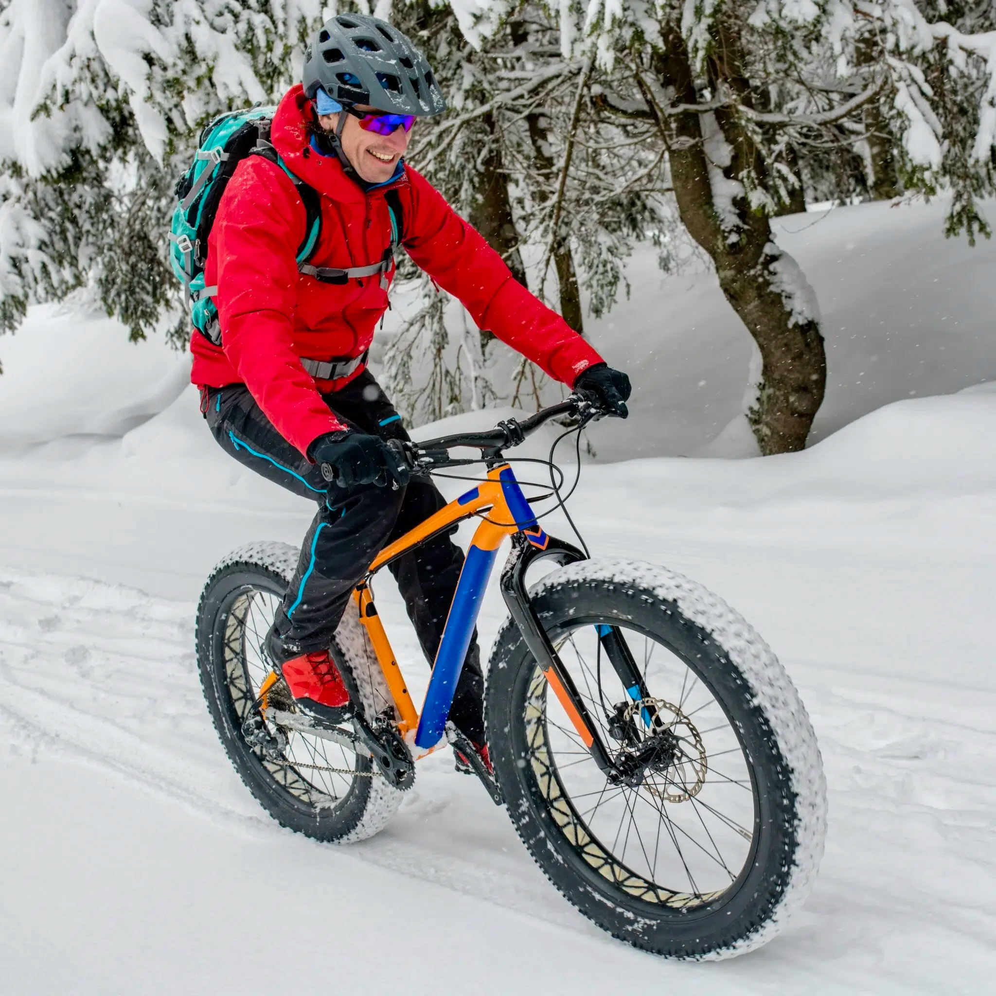 Man riding a fat tire mountain bike on snowy trail, wearing red jacket, helmet, and sunglasses, with snow-covered trees in the background.