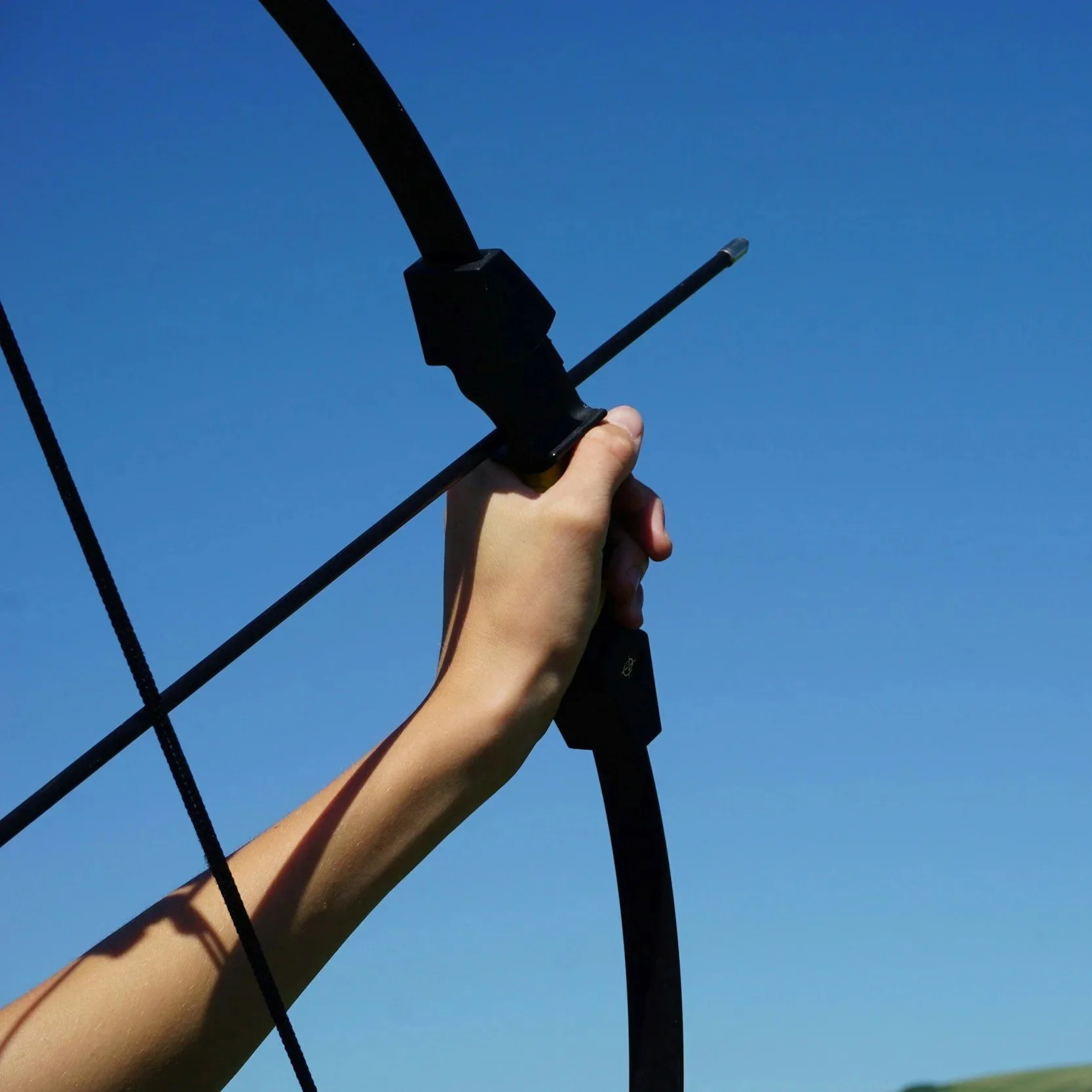 Close-up of a hand holding a black bow with an arrow ready to shoot against a clear blue sky.