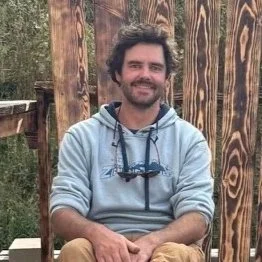 A man with dark, curly hair and a beard smiling while sitting outside in front of a wooden fence, wearing a light gray hoodie.