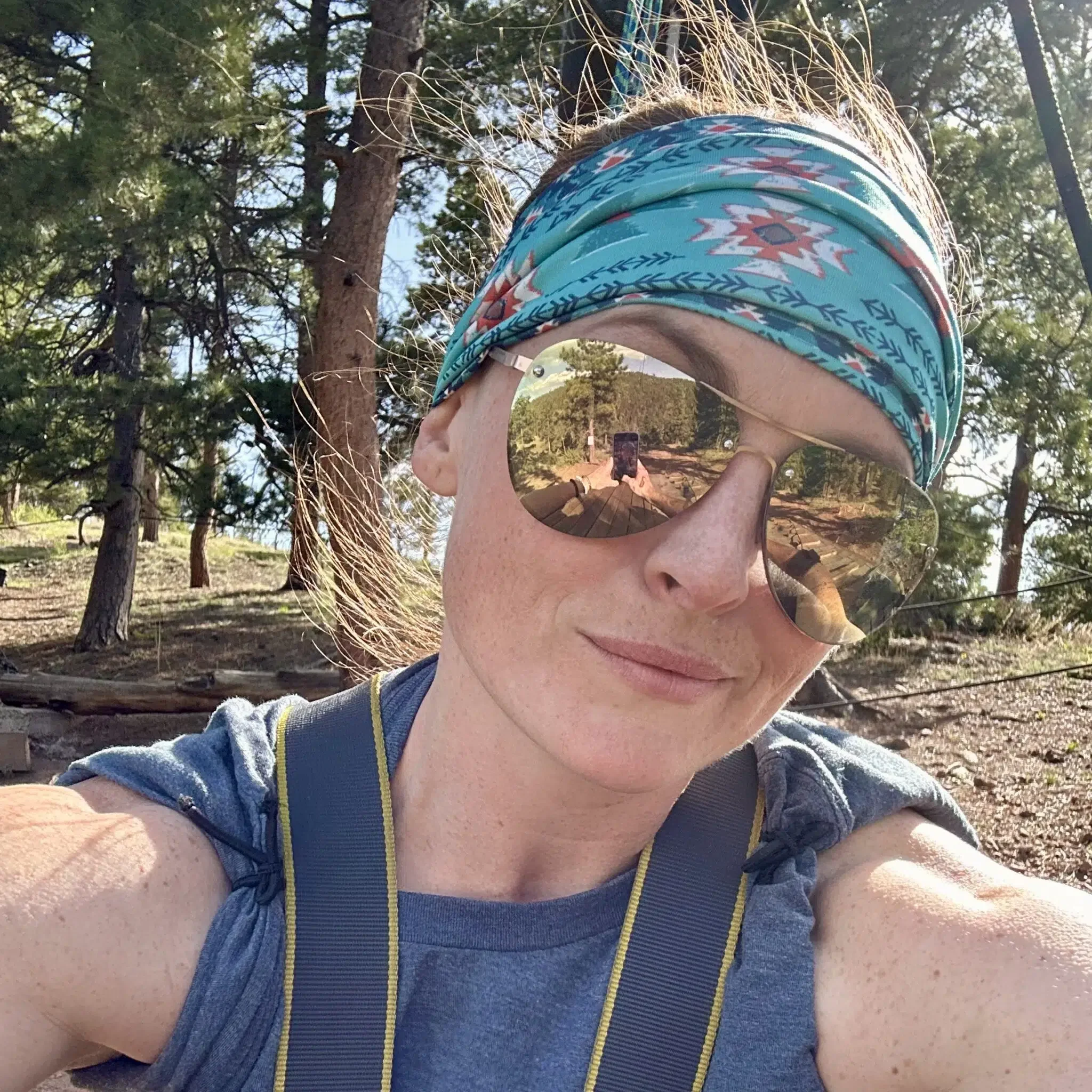 A woman taking a selfie outdoors in a forested area, wearing sunglasses, a colorful headband, and a blue sleeveless top.