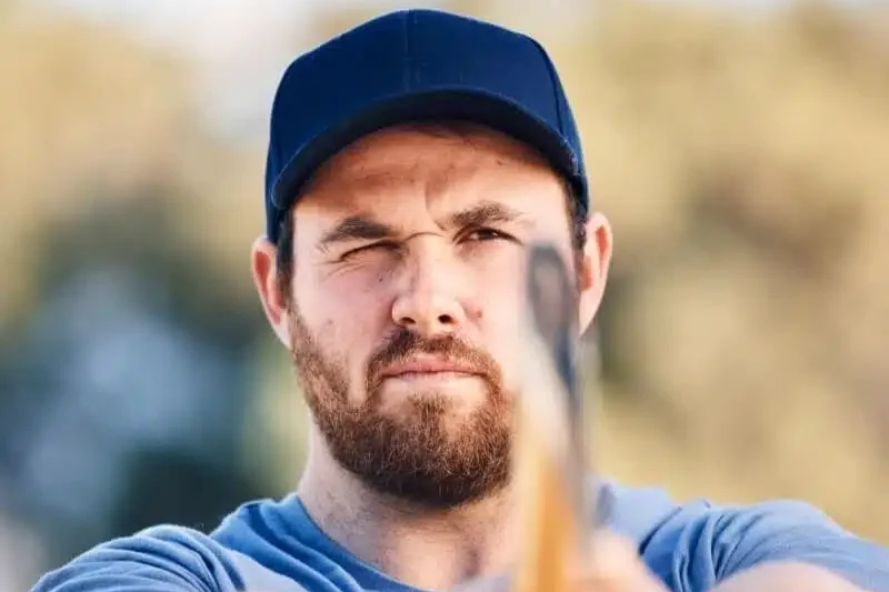 A man with a beard and mustache wearing a blue cap and blue shirt, aiming a bow and arrow with a serious expression.