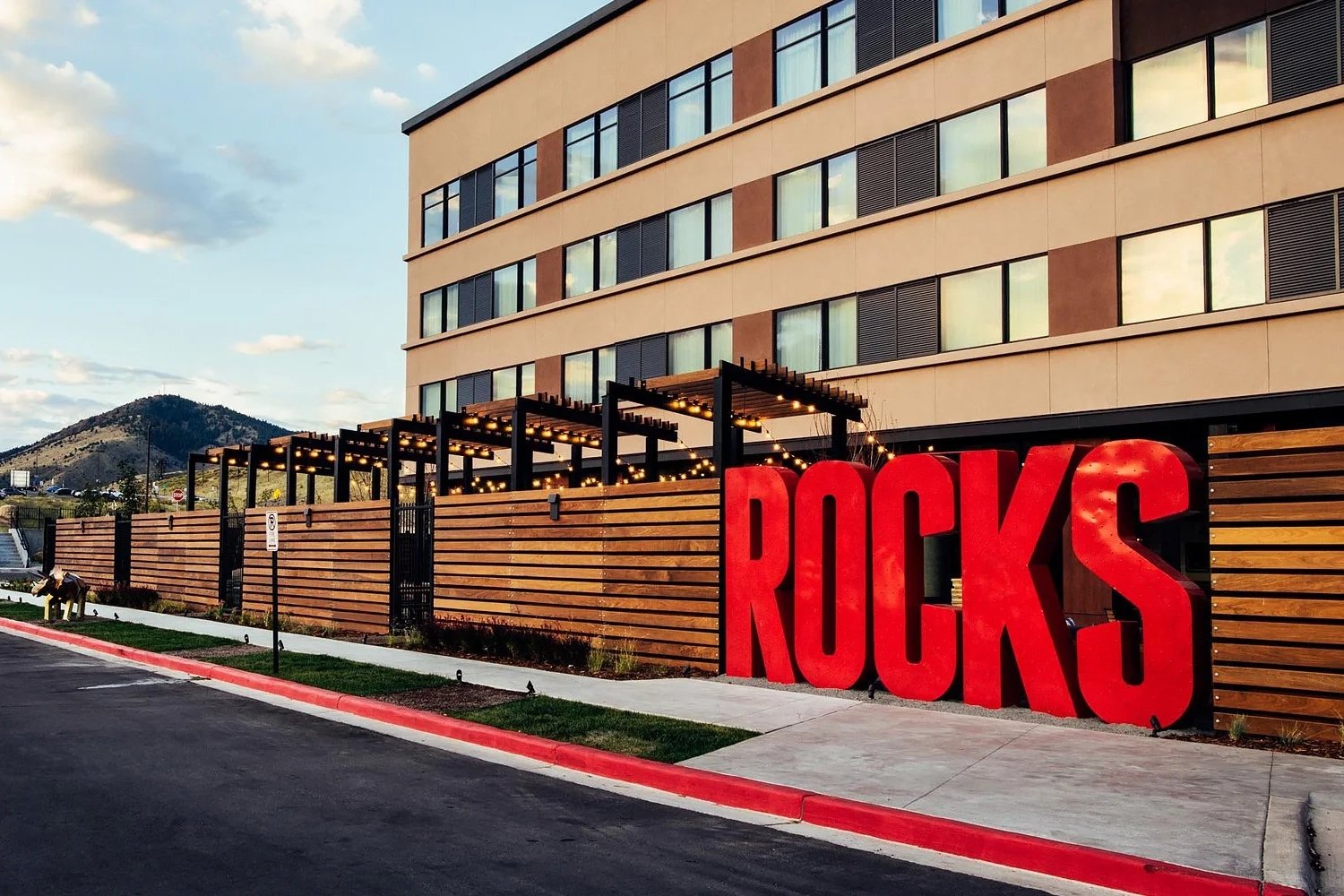 Red large letters spelling 'ROCKS' next to a wooden fence outside a modern multi-story building with mountains in the background.