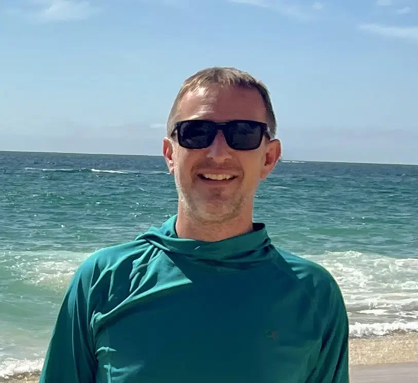 A man wearing sunglasses smiling at the beach with the ocean and blue sky in the background.