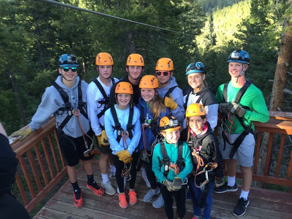 Group of children and teens in safety gear, including helmets and harnesses, standing on a wooden platform outdoors.