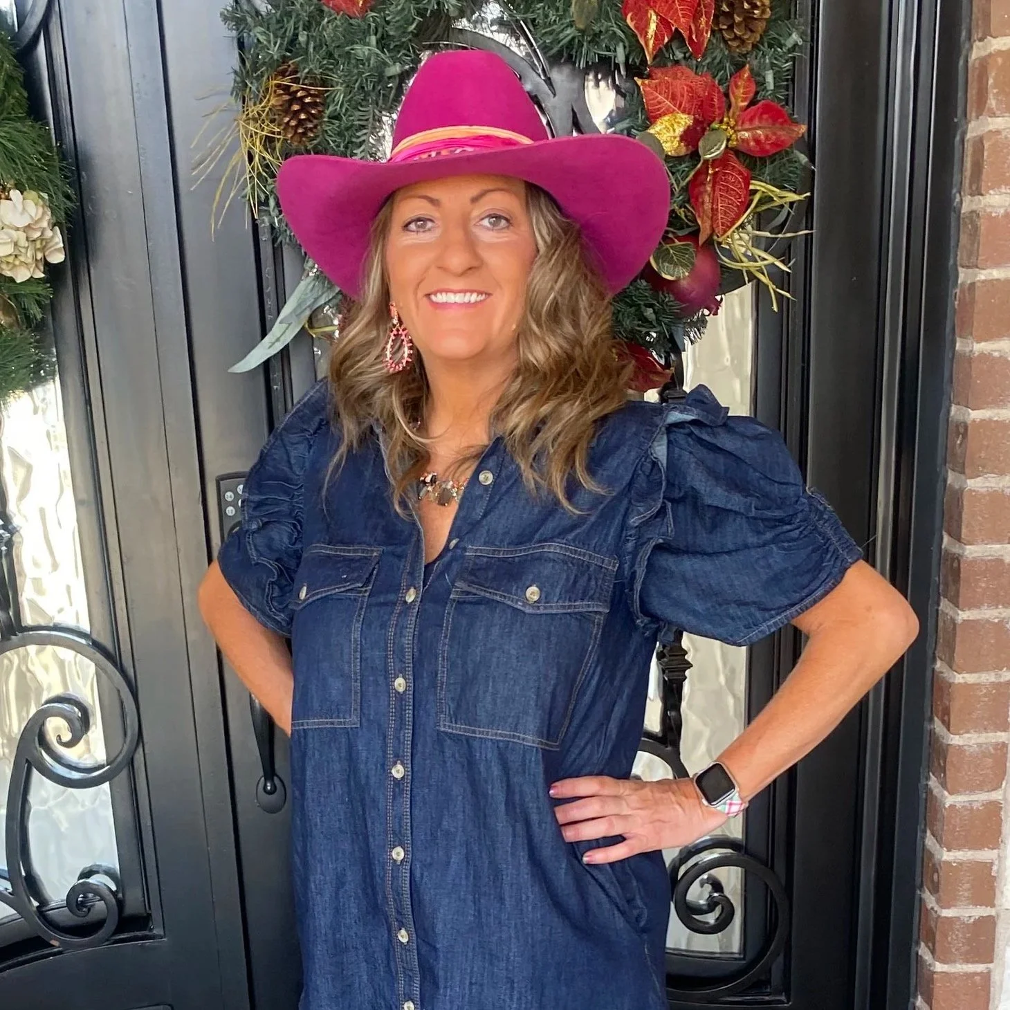 Woman wearing a large pink hat, denim dress, and jewelry, standing in front of holiday wreath decoration.