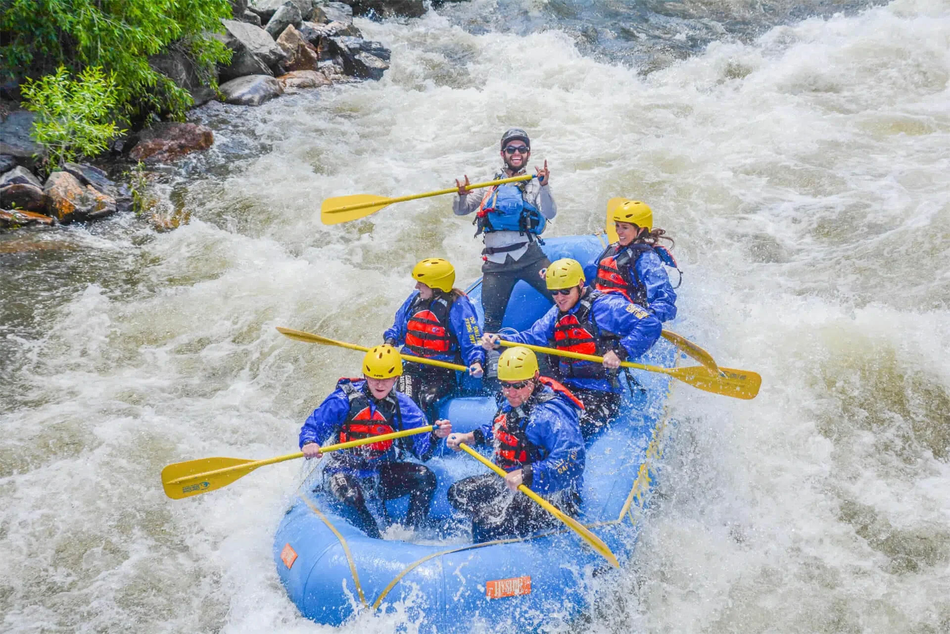 A group of six people white water rafting in a river, wearing blue jackets, yellow helmets, and red life vests, with a guide at the back paddling and giving signals.