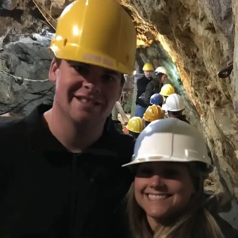 Two people wearing safety helmets, one yellow and one white, in a cave or mine with other workers in the background.