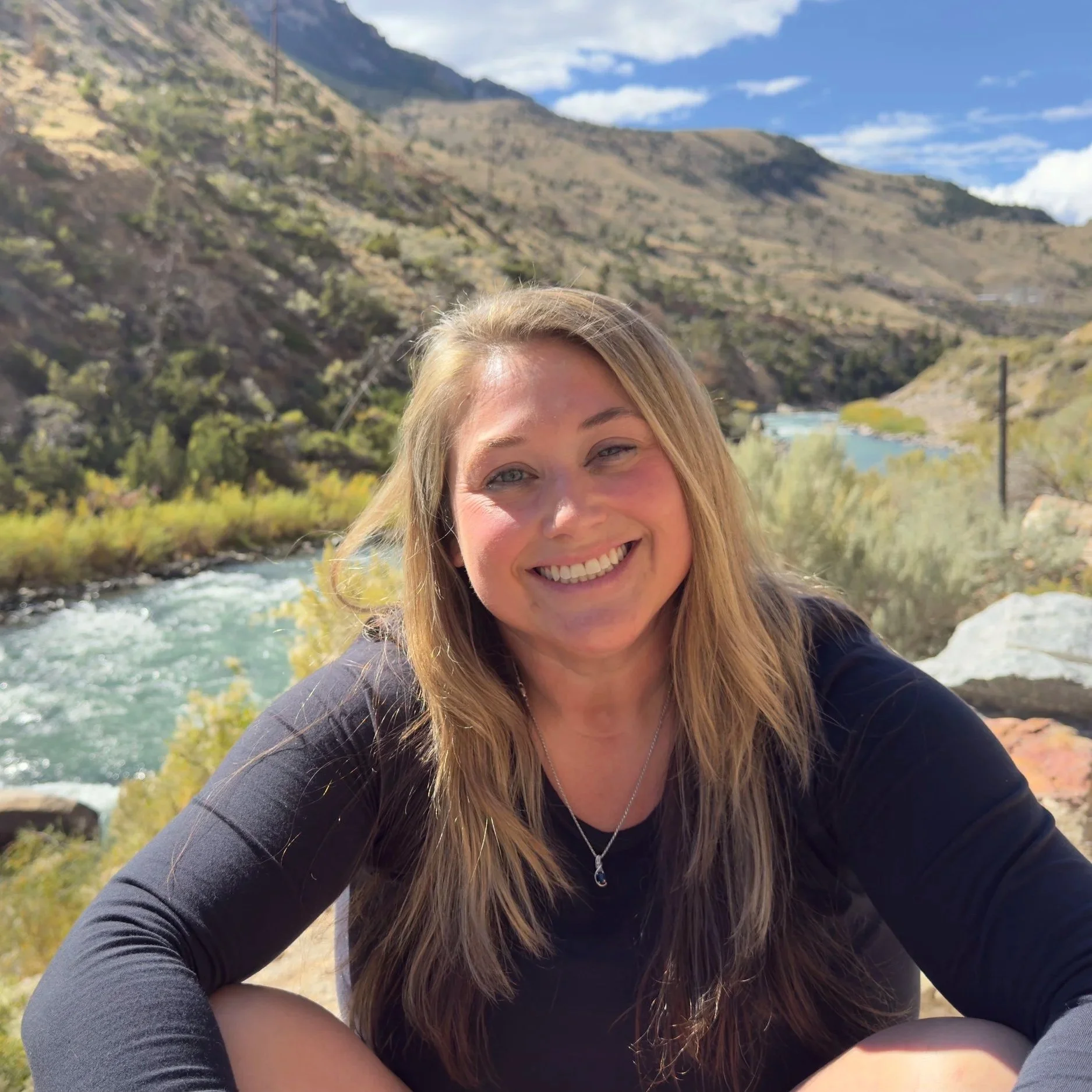 A woman smiling outdoors in a mountainous landscape near a river.