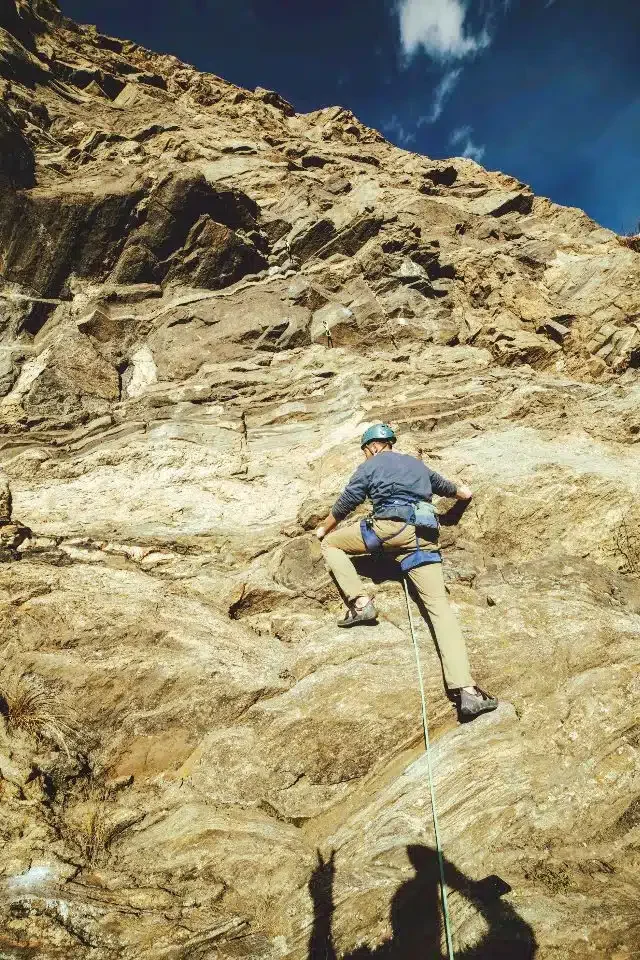 A person in climbing gear ascending a rocky cliff face outdoors under a blue sky.