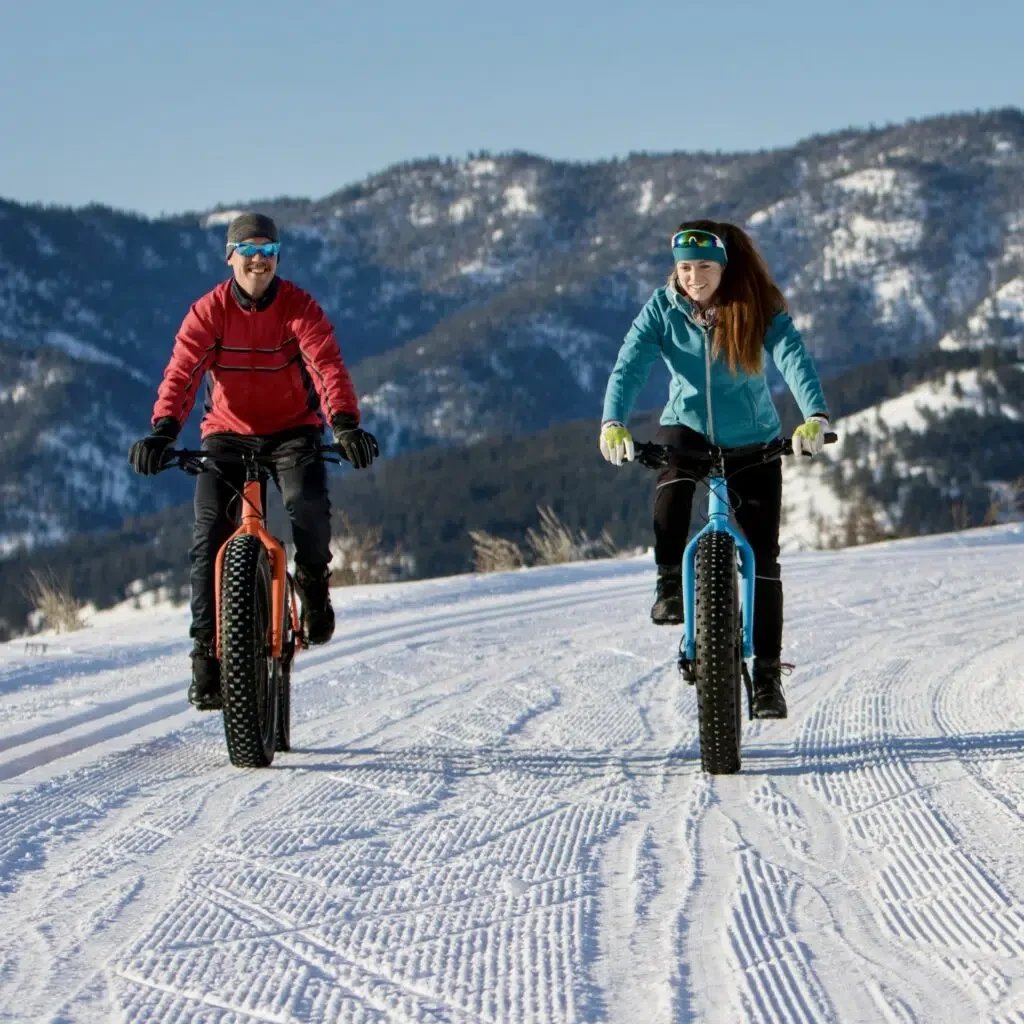 Two people riding fat tire bikes on a snowy trail in a mountainous area with snow-covered hills in the background.