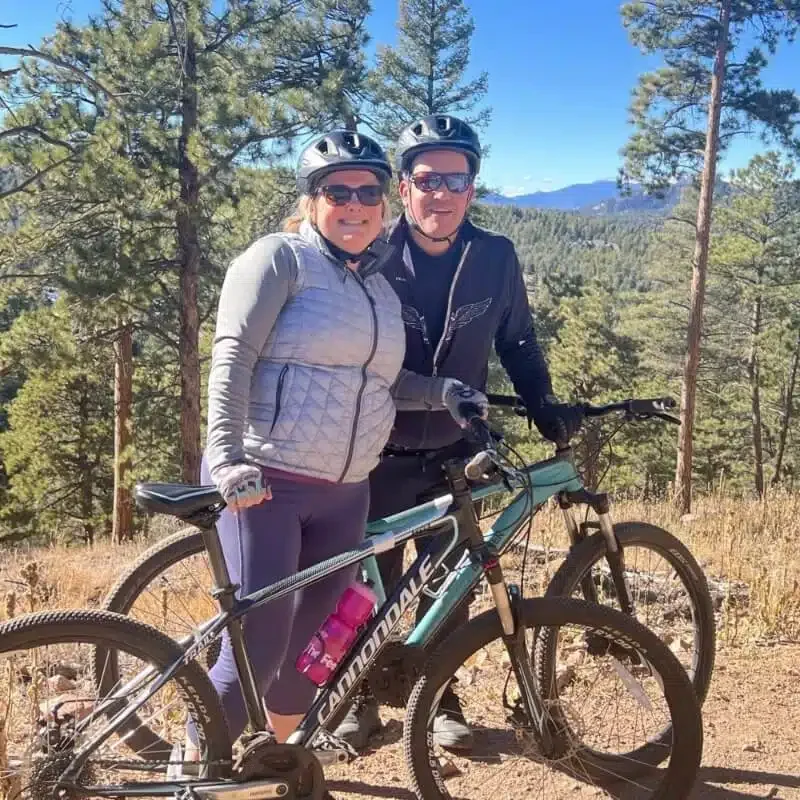 A couple wearing helmets and sunglasses standing next to a mountain bike in a forested area with mountains in the background.