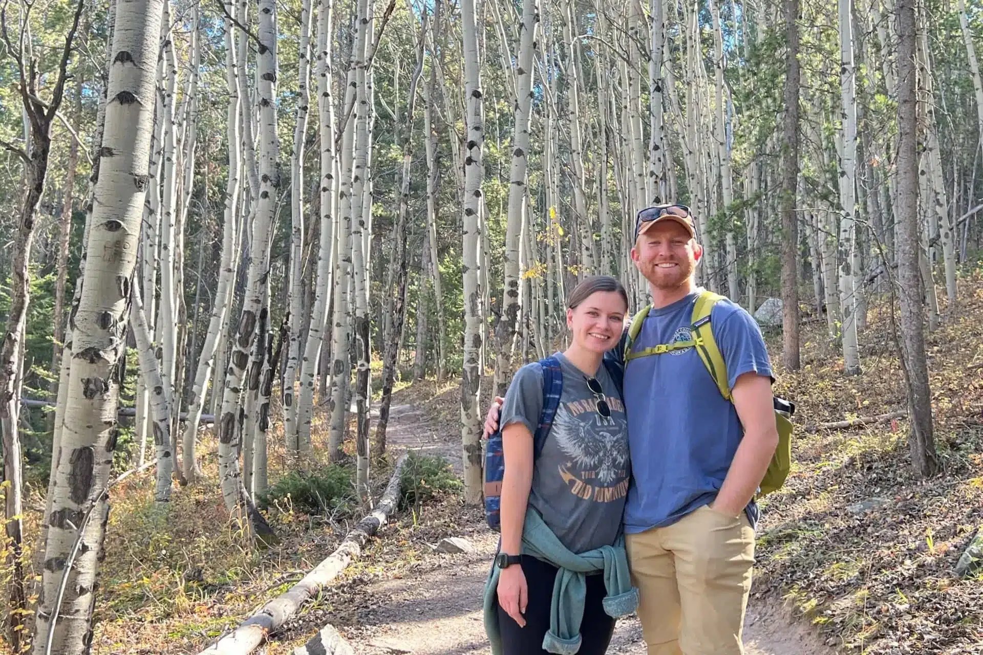 A smiling couple hiking in a forest of tall white-barked aspen trees with green foliage in the background.