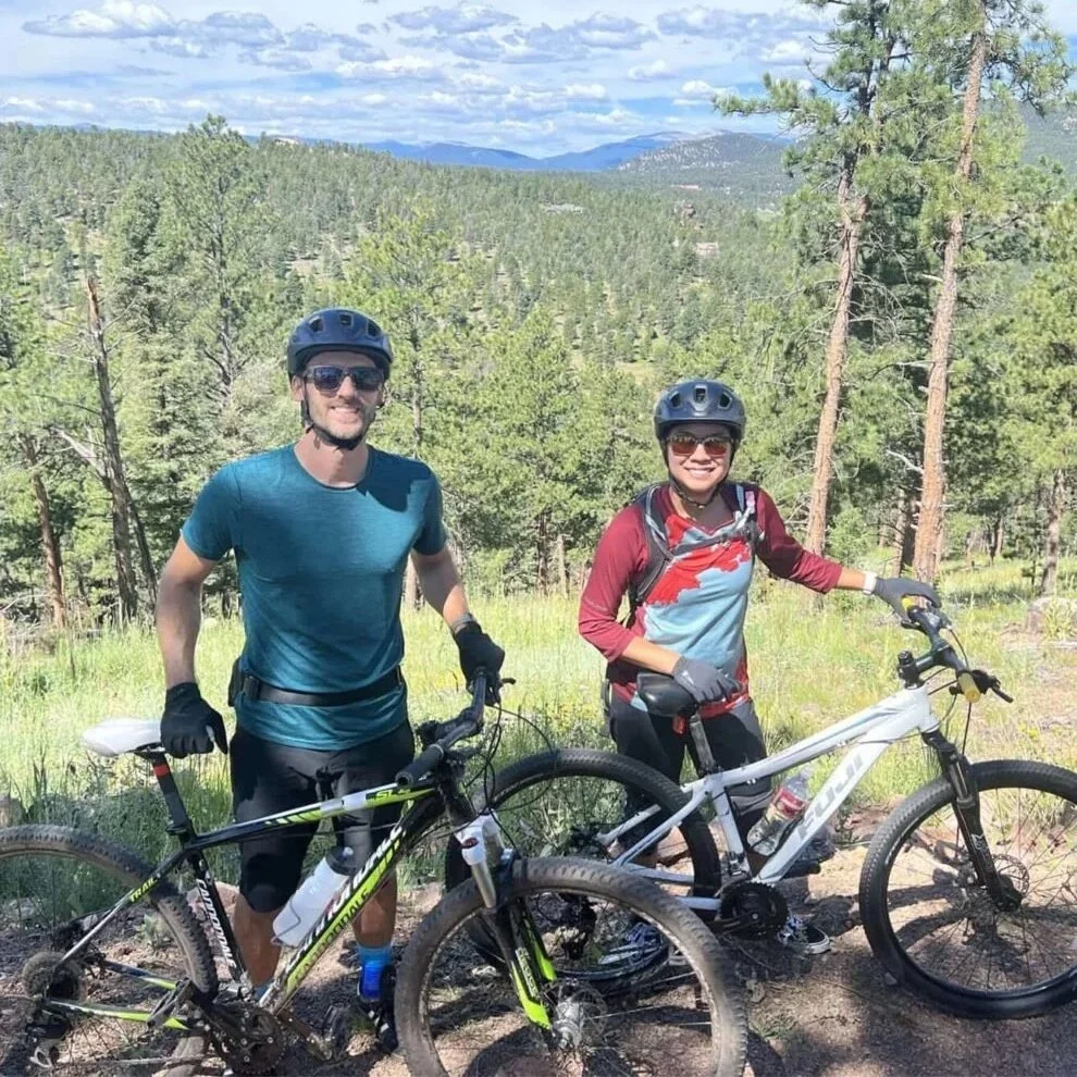 Two people wearing helmets and sunglasses standing with mountain bikes on a forested trail in a mountainous area. The background shows dense trees, green hills, and a partly cloudy sky.