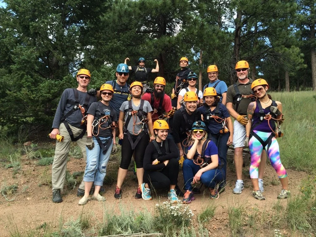 Group of people wearing safety helmets and harnesses, posing outdoors on a dirt trail surrounded by trees.