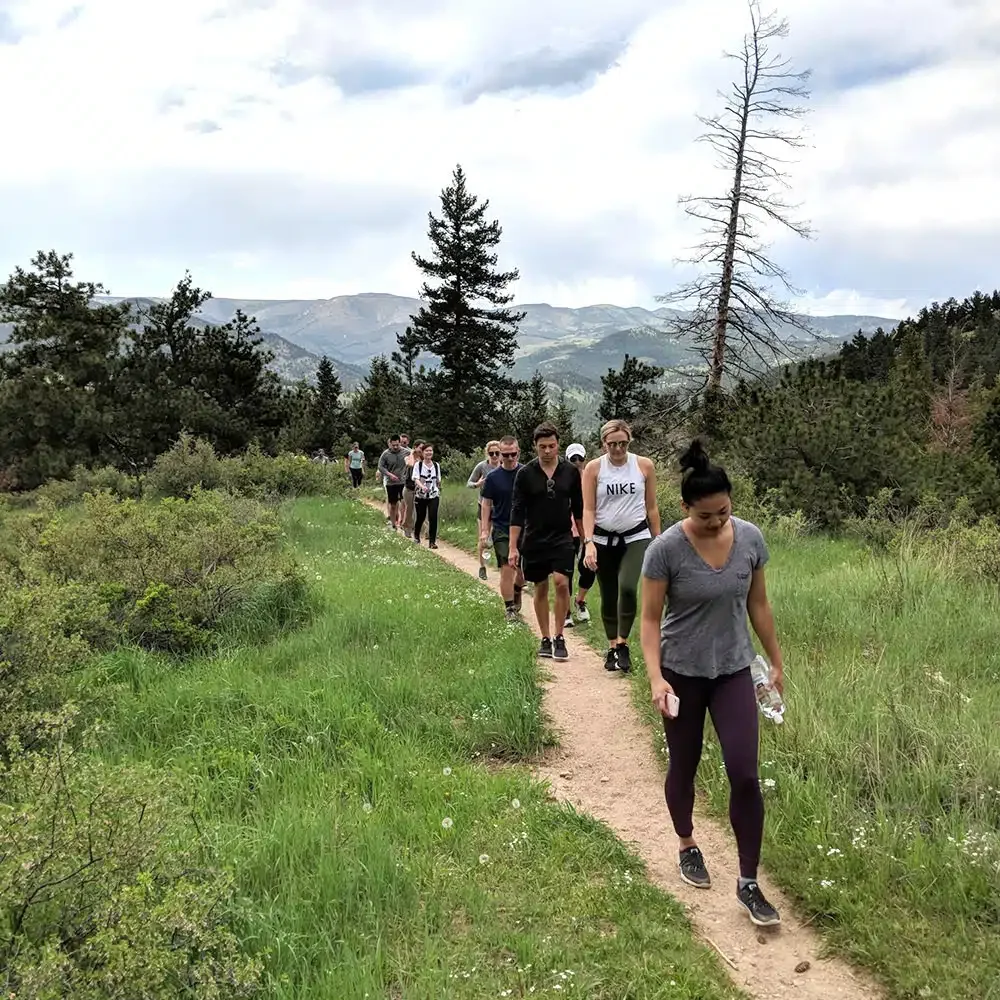 People hiking on a narrow dirt trail through a green mountain landscape with trees and distant mountains under a cloudy sky.
