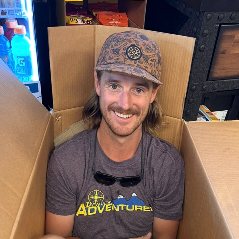 Man smiling while sitting inside a large cardboard box, wearing a cap and glasses around neck, in a store setting.