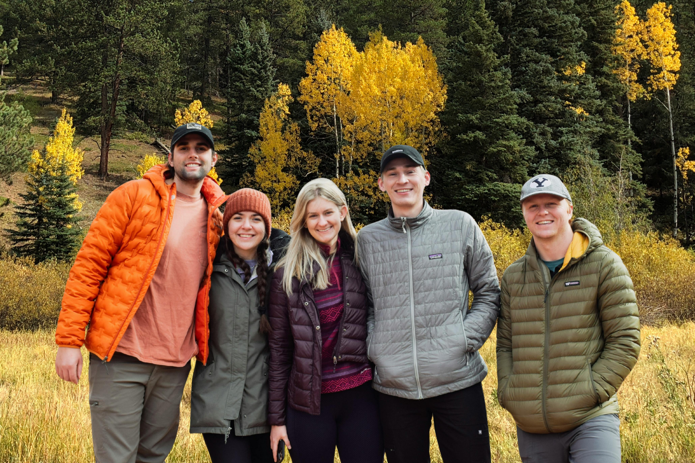 Group of five young adults outdoors in fall, standing in a grassy field with trees showing yellow and green leaves in the background, smiling at the camera.